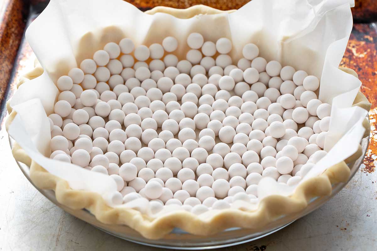 Small white, round ceramic pie weights fill a raw pie crust lined with a sheet or parchment paper.