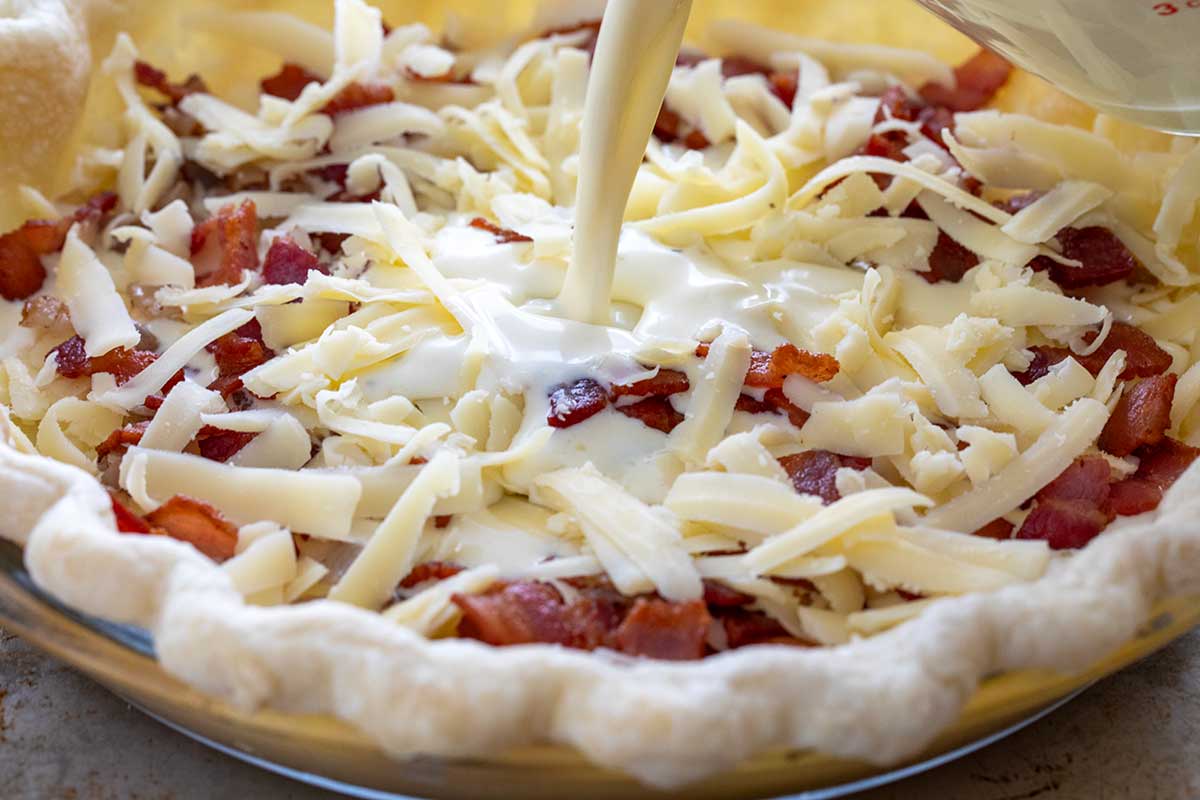 Pouring lorraine filling into a pie crust filled with bacon and cheese.