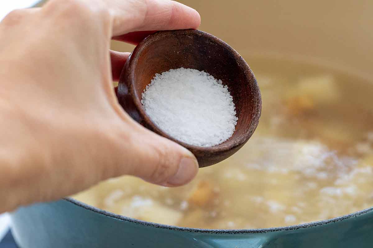 A hand holding a small wooden bowl of coarse salt over a pot of cubed potatoes in boiling water.