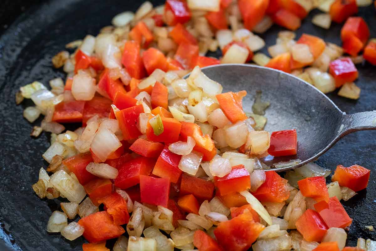 Diced onions and red bell peppers sautéing in a dark skillet with a metal spoon.