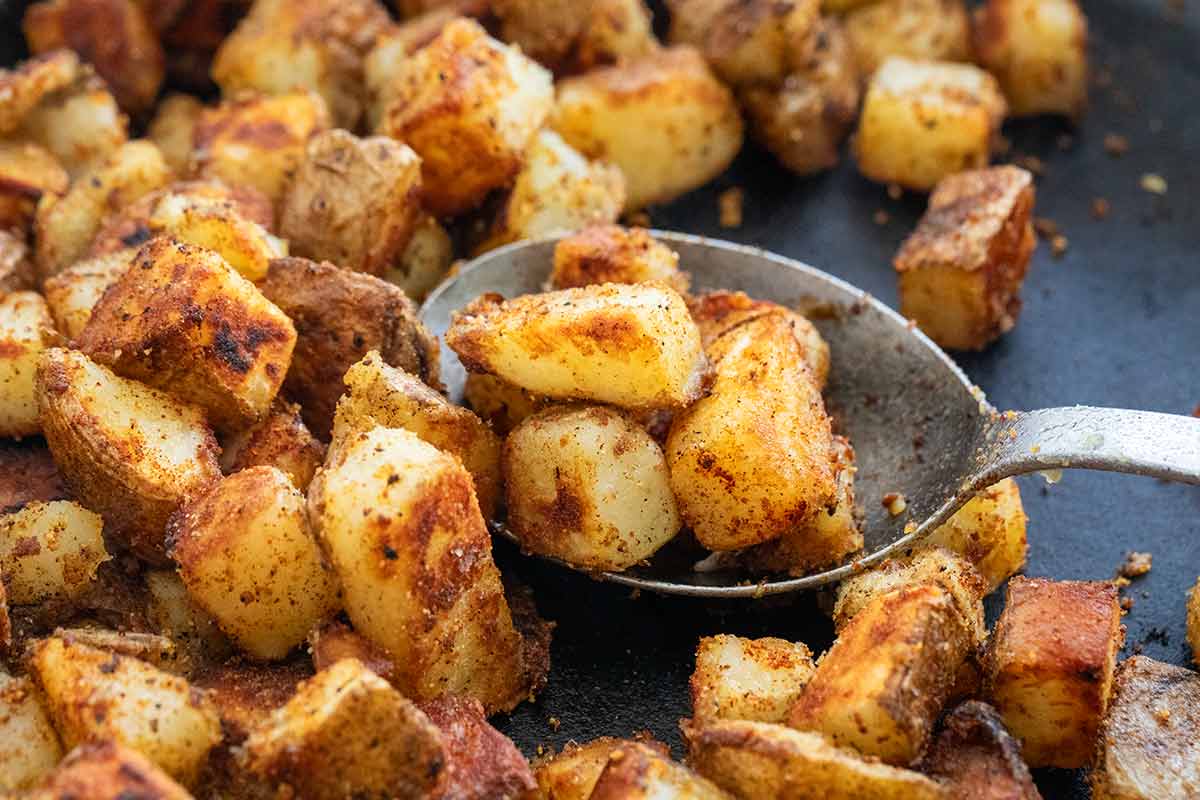 A spoon scoops up crisp, golden-brown diced potatoes in a skillet.
