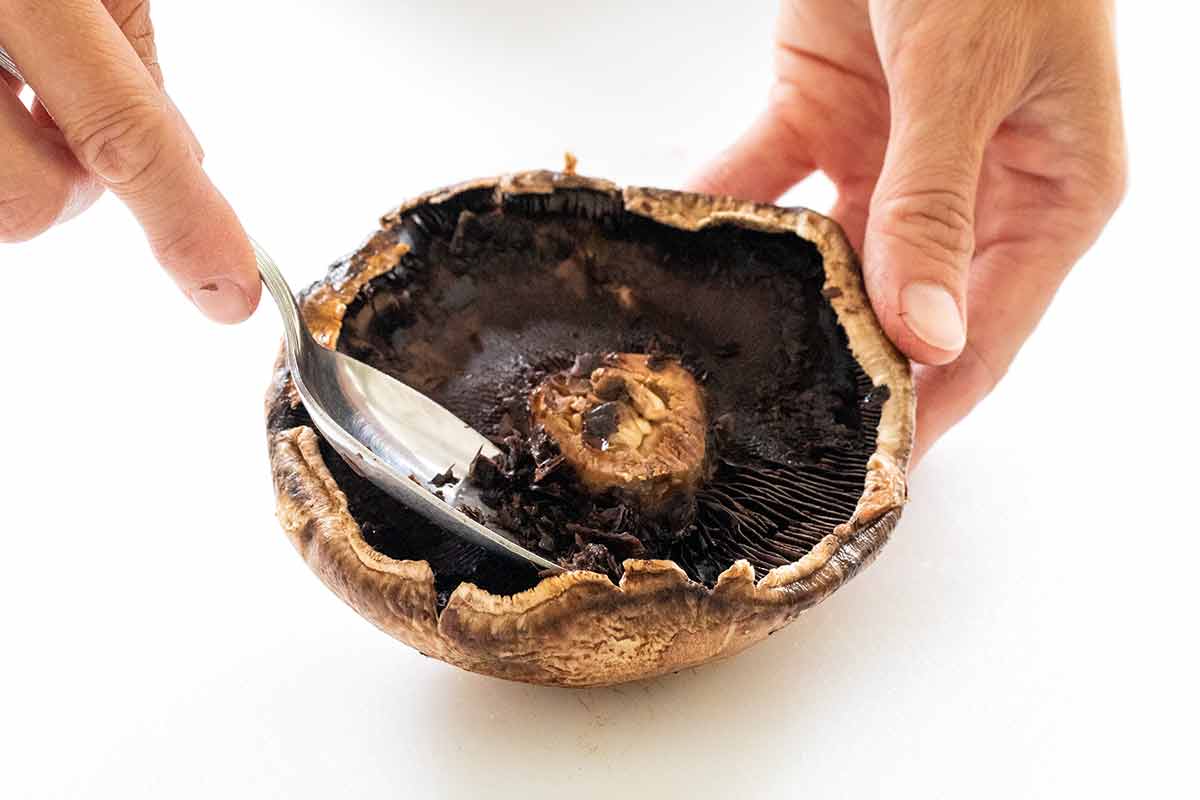 Person using a metal spoon to scoop out the gills of a portobello mushroom cap.