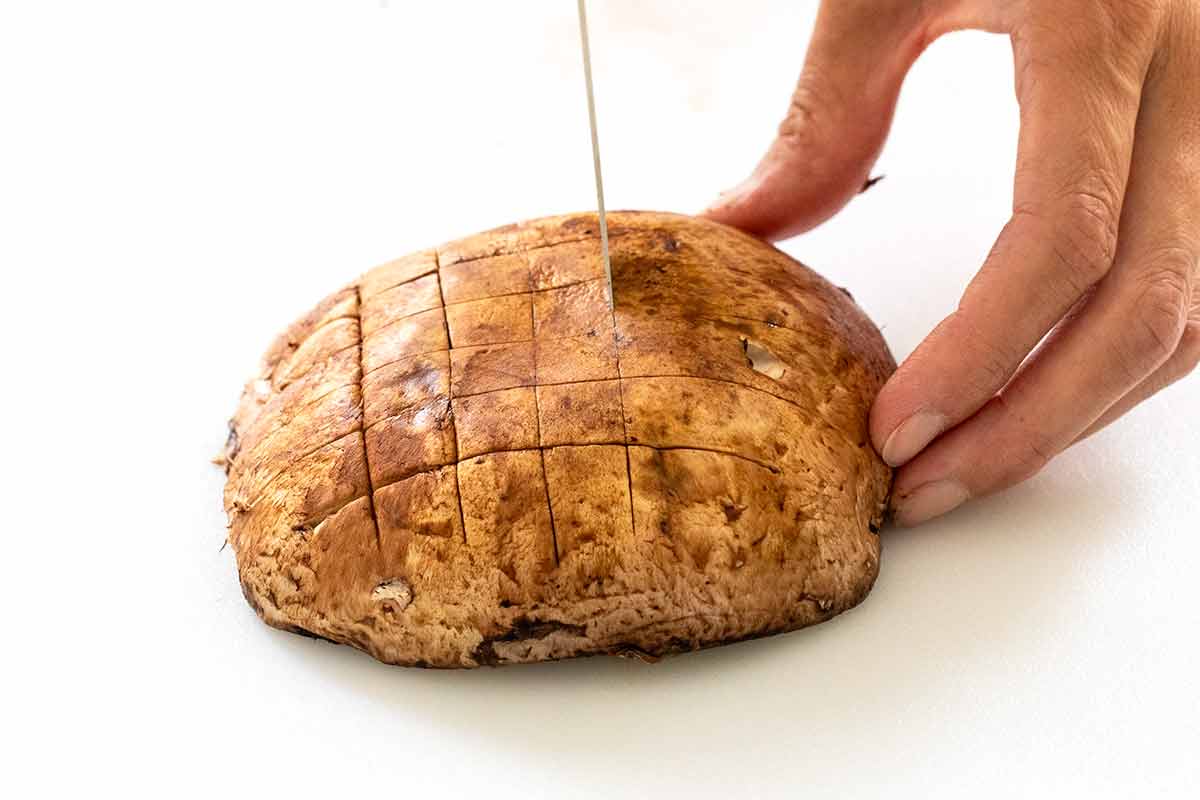 Person holding the side of a portobello mushroom and making a crosshatch knife marks on top.