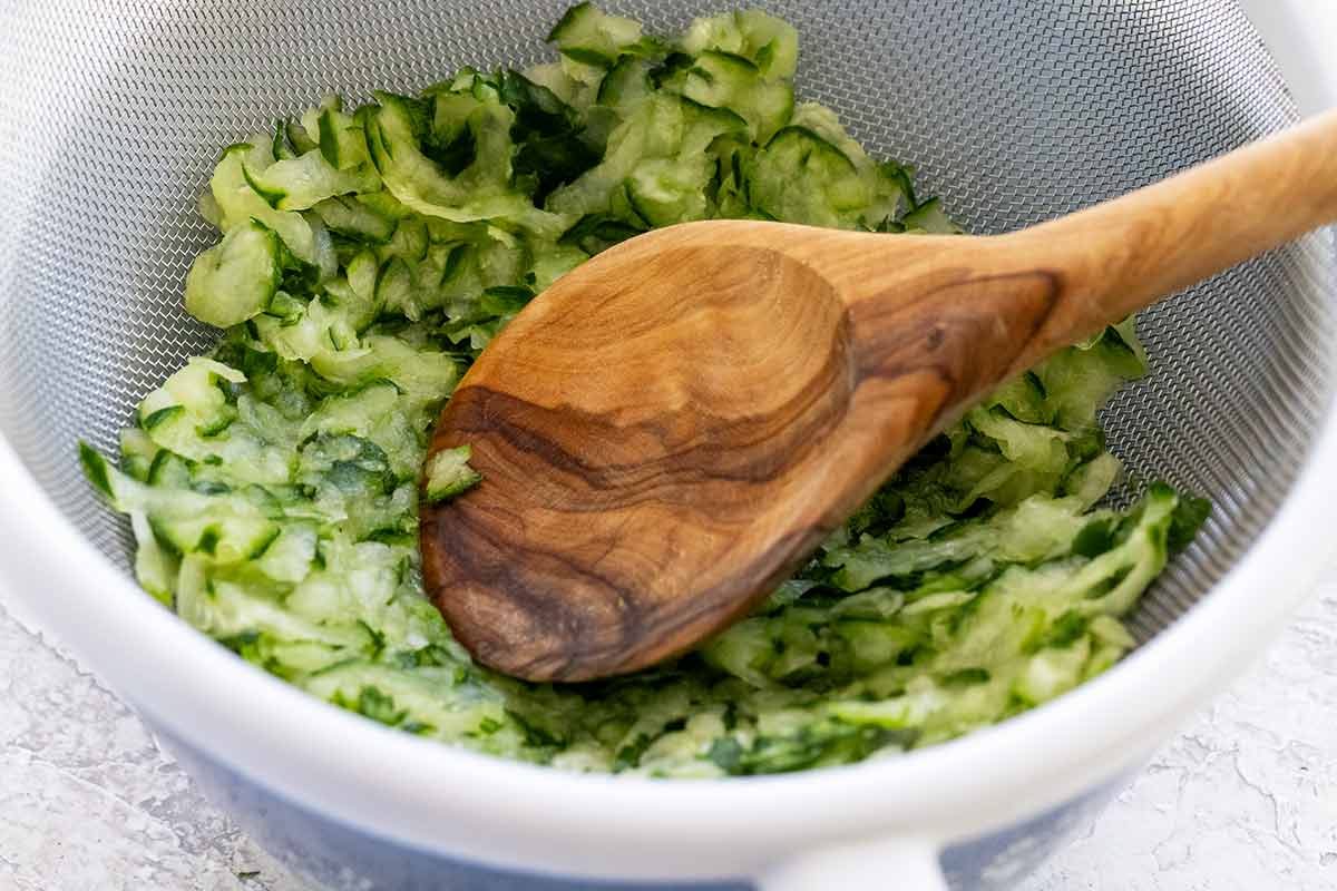 Wooden spoon pressing shreds of cucumber into a mesh stainer.