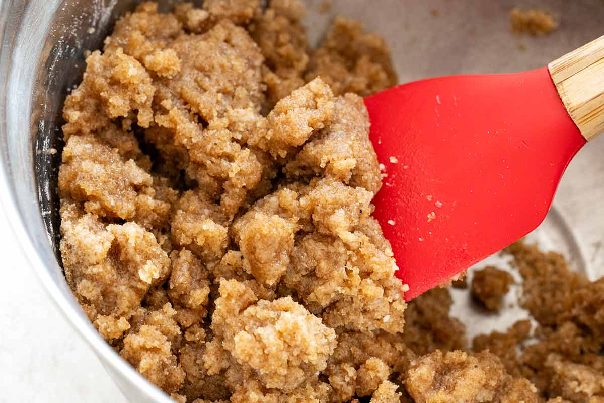 Clumpy brown crumble topping mixture being stirred in a metal bowl with a red spatula.