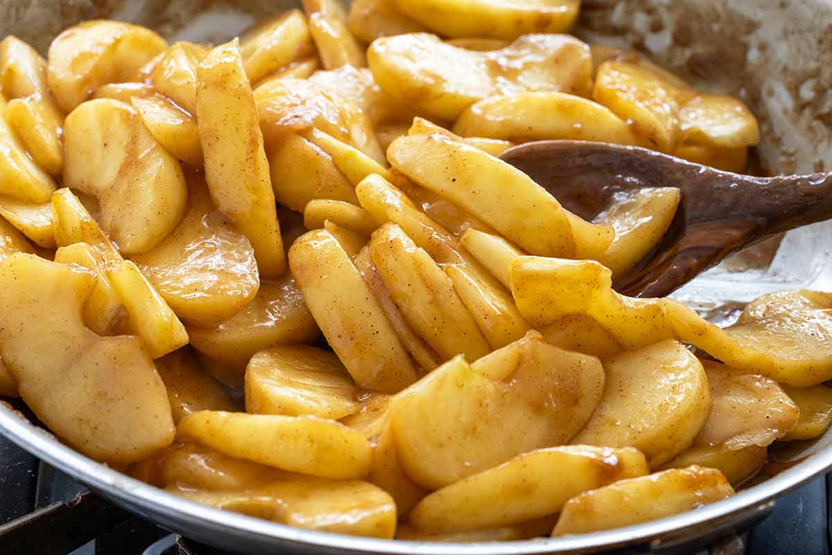 Sliced apples coated in spiced brown sugar syrup being stirred with a wooden spoon in a pan.