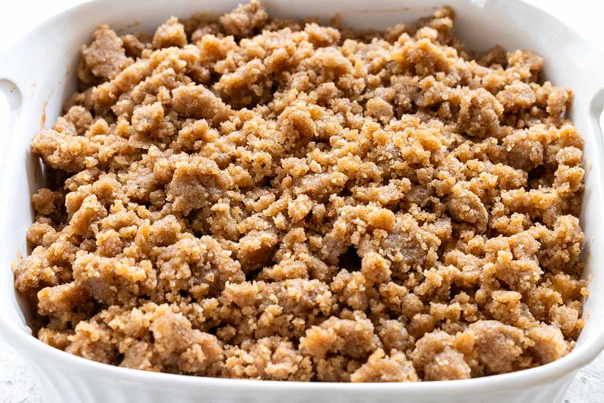 Crumble topping spread in an even layer over cooked apples in a white baking dish.