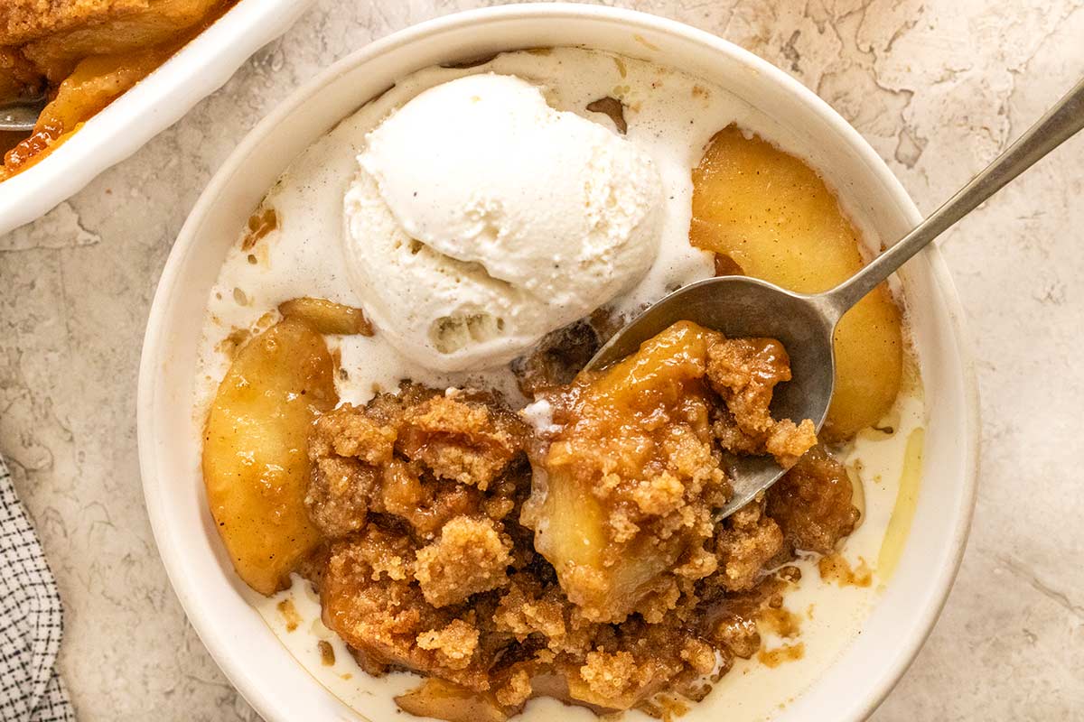 Bowl of warm apple crumble topped with melting vanilla ice cream, with a spoon taking a bite.
