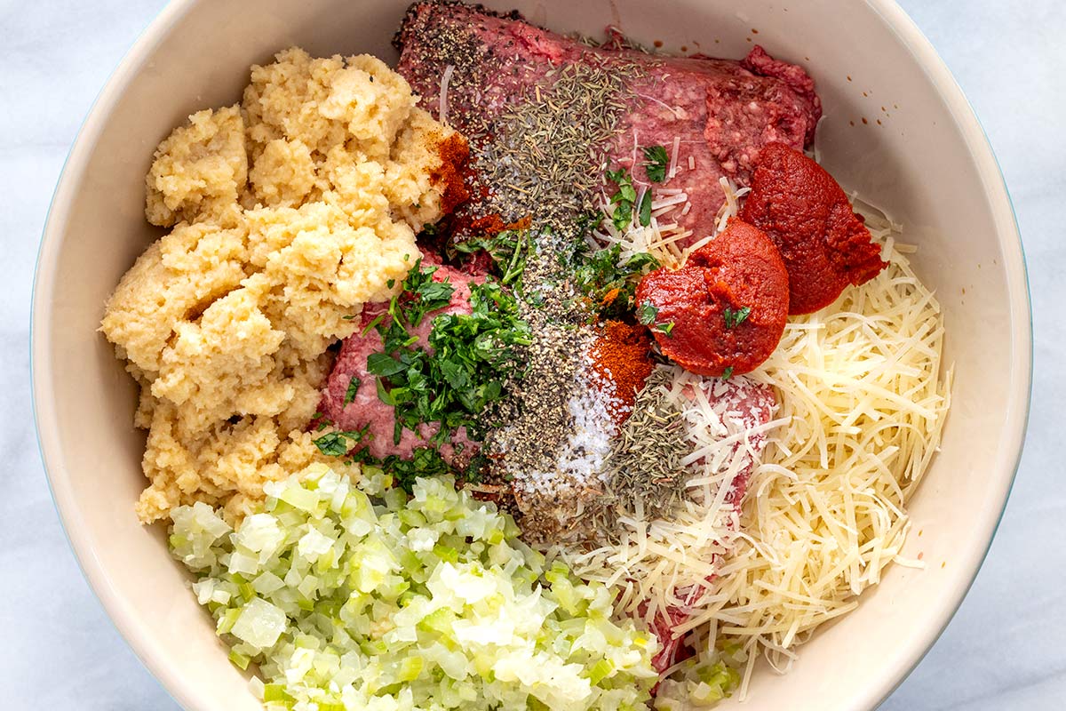 Raw ground beef, panade, sautéed vegetables, parmesan cheese, tomato paste, and spices in a large mixing bowl before mixing.