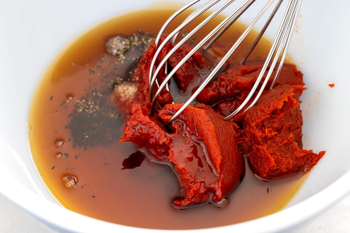 Tomato paste, liquid glaze ingredients, and black pepper in a white bowl with a wire whisk.