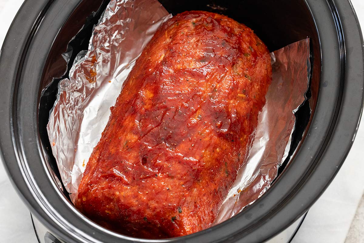 Glazed raw meatloaf resting on a foil sling inside a slow cooker pot.