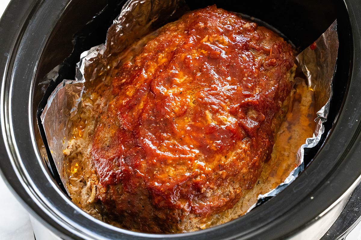 Glazed meatloaf resting on an aluminum foil sling inside a slow cooker insert.