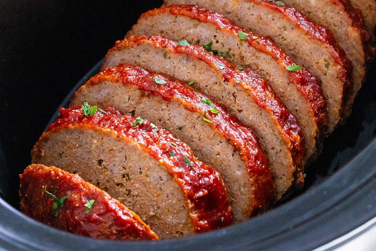 Sliced meatloaf topped with red glaze and chopped herbs inside a slow cooker.