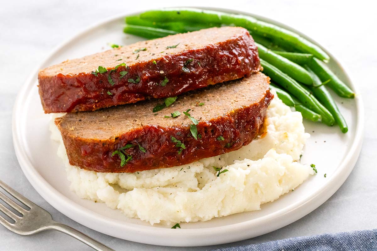 Two thick slices of glazed meatloaf topped with chopped herbs, served over mashed potatoes with green beans on the side.