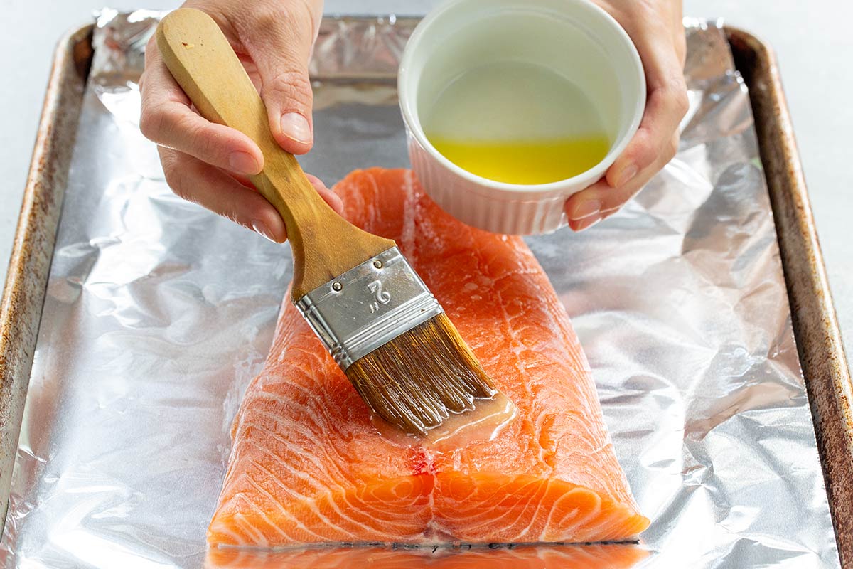 Person brushing melted butter on top of a salmon fillet placed on a foil lined sheet pan.