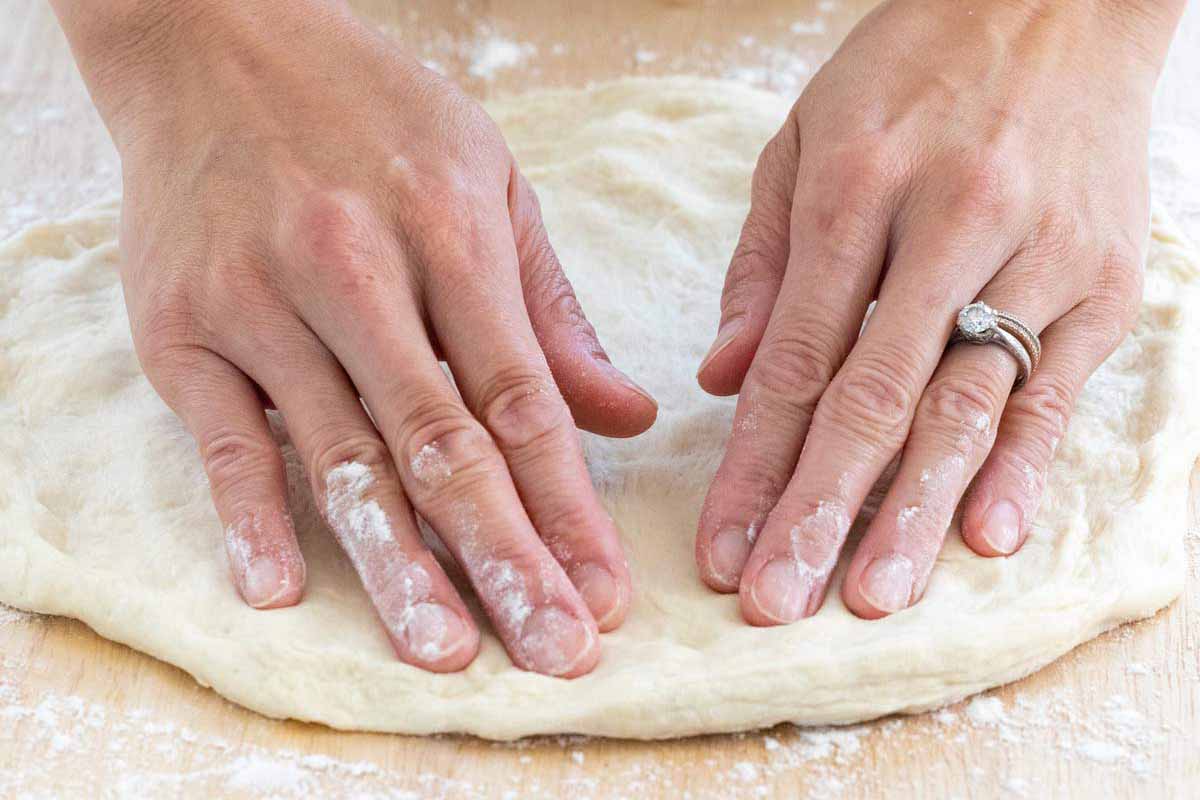 Persons hands shaping a raw pizza dough on a floured surface.