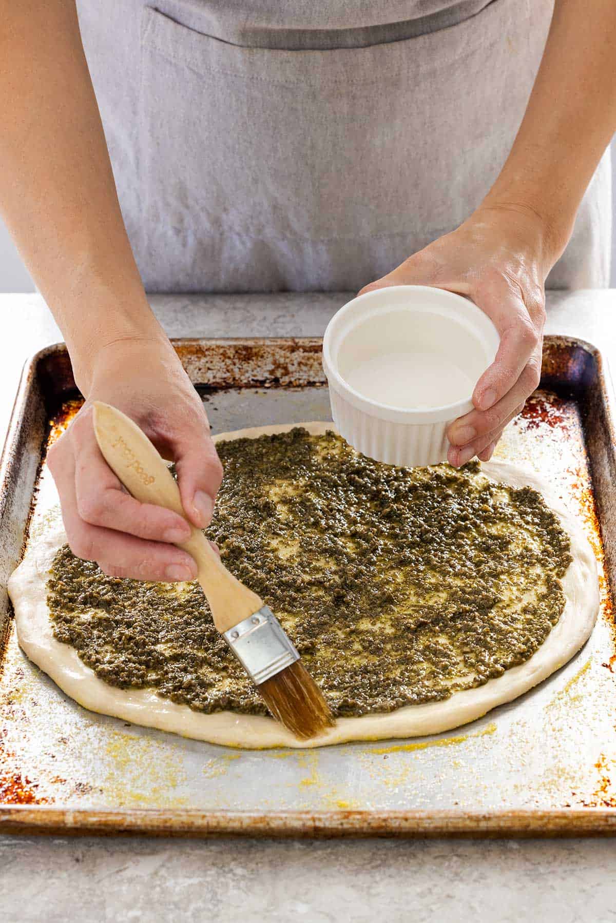 Olive oil being brushed on the edge of a pizza crust with pesto sauce.