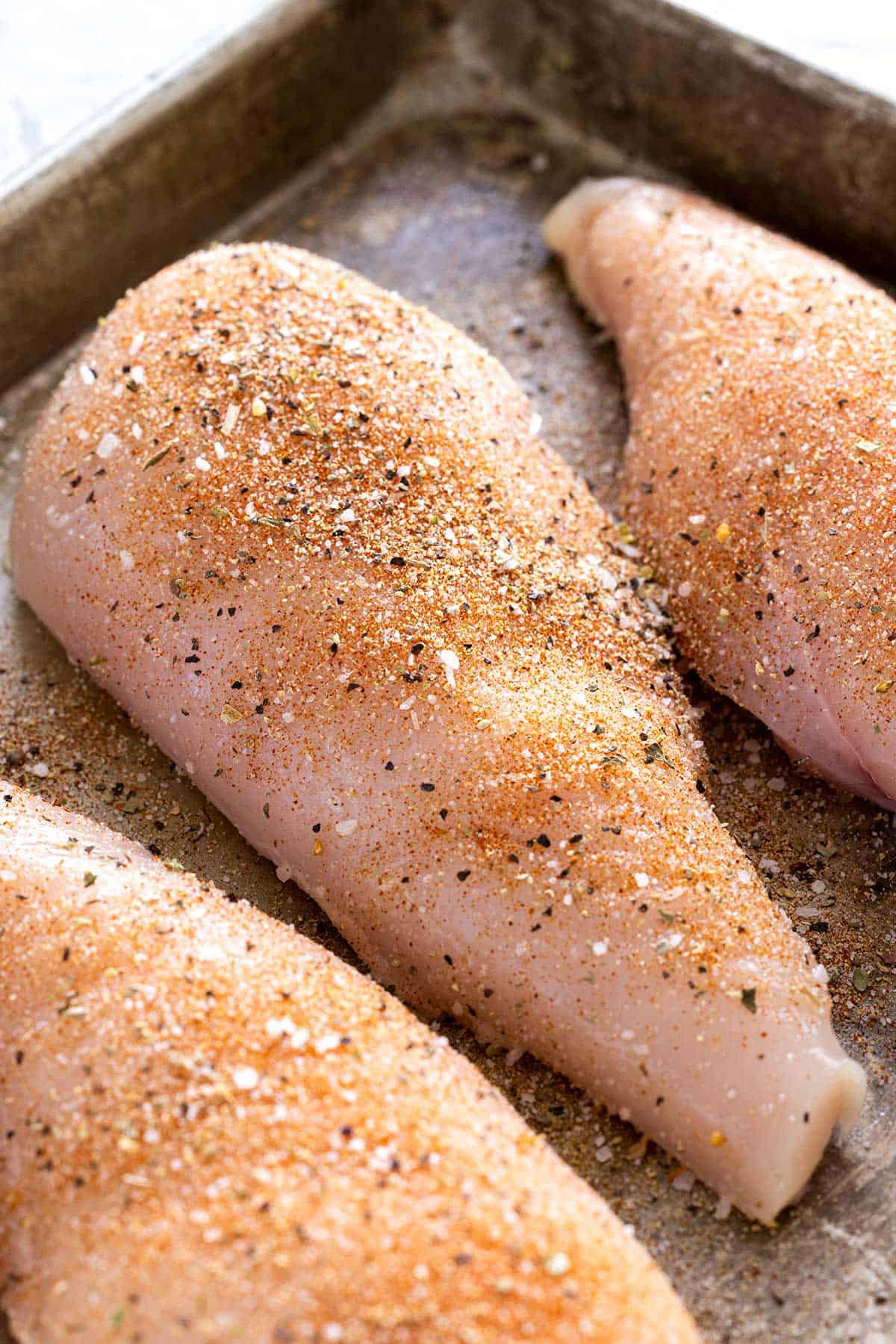 Sheet pan with raw chicken breasts that have dried seasonings on their surface.