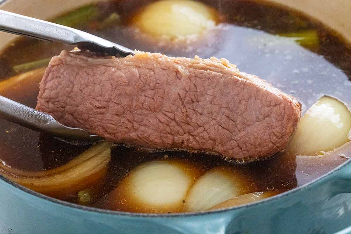 Tongs removing a brisket from the cooking liquid.