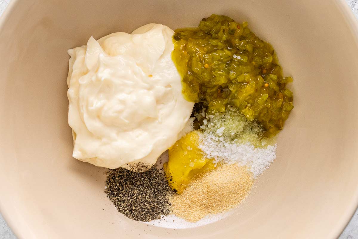Dressing ingredients including mayonnaise, sweet relish, yellow mustard, and spices placed in a mixing bowl.