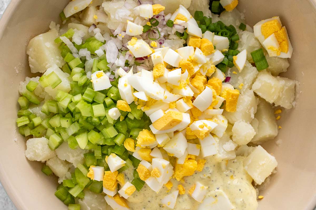 Diced potatoes, chopped hard-boiled eggs, celery, red and green onions, and mayonnaise dressing in a large bowl.