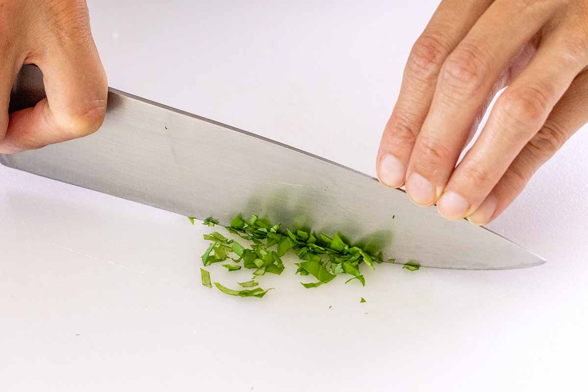 Person rocking a chefs knife to chop basil on a white cutting board.