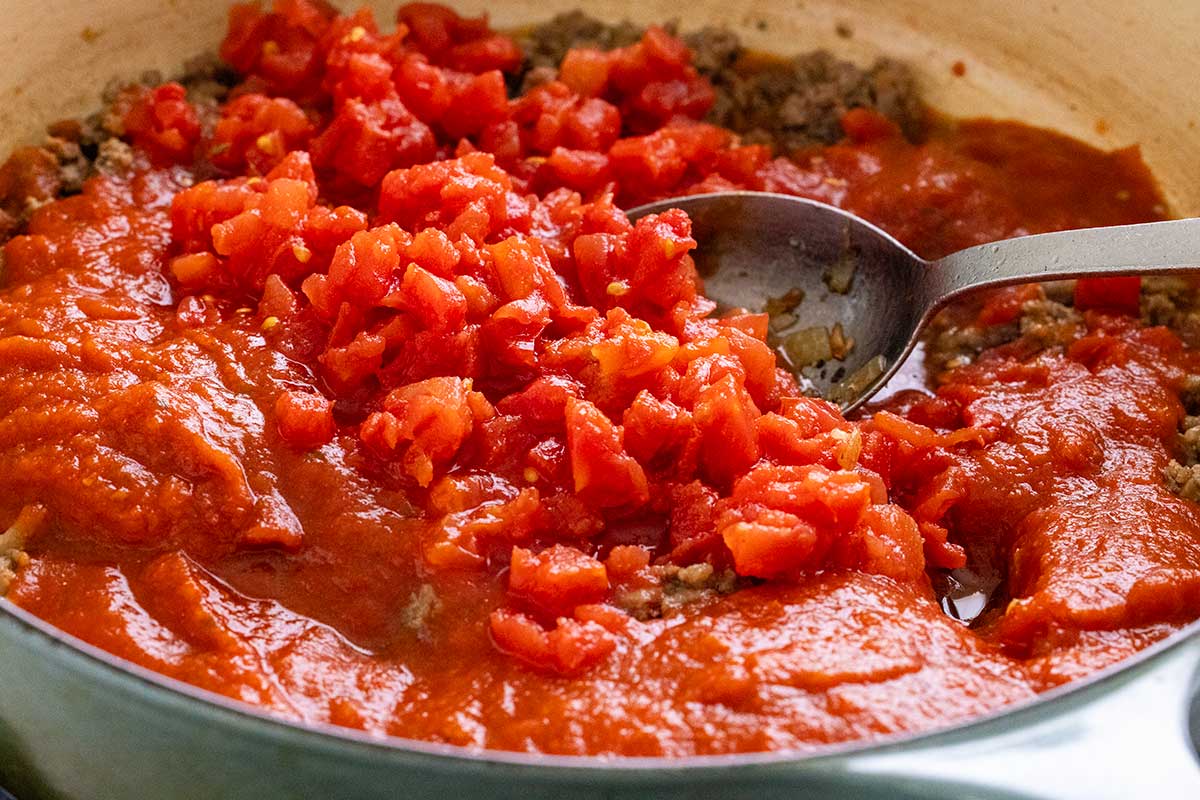 Diced tomatoes and marinara sauce poured over browned meat in a large pot.