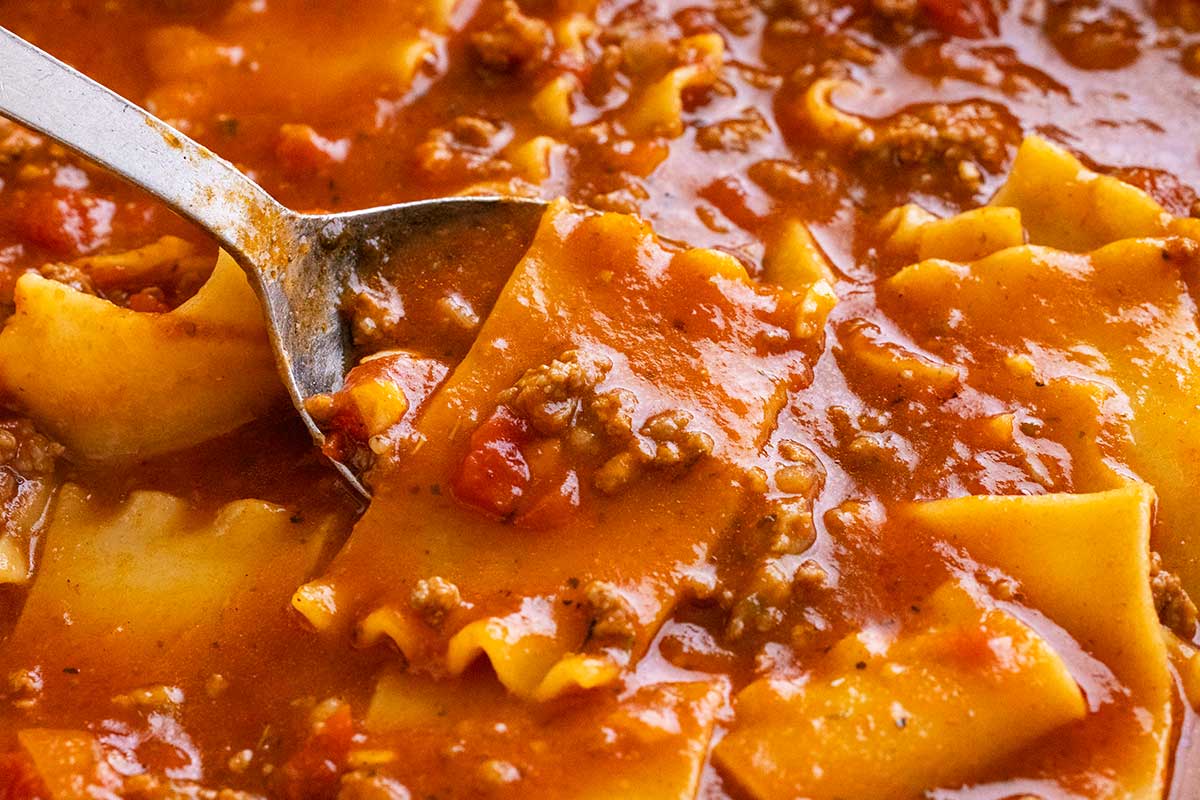 A metal spoon scoops broken lasagna noodles simmering in a rich tomato and ground meat soup broth.