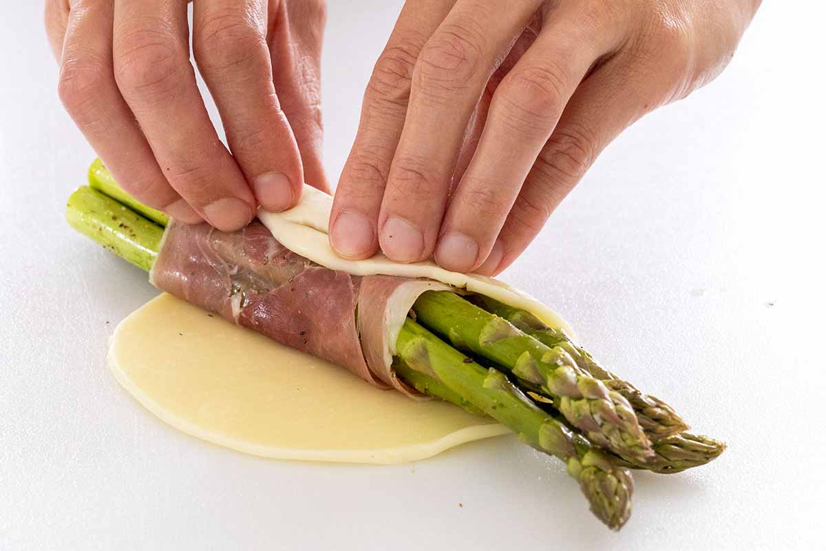 Person rolling cheese and prosciutto around a bundle of asparagus.