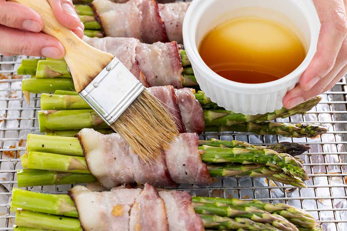Maple syrup being brushed on strips of bacon wrapped around a bunch of asparagus.