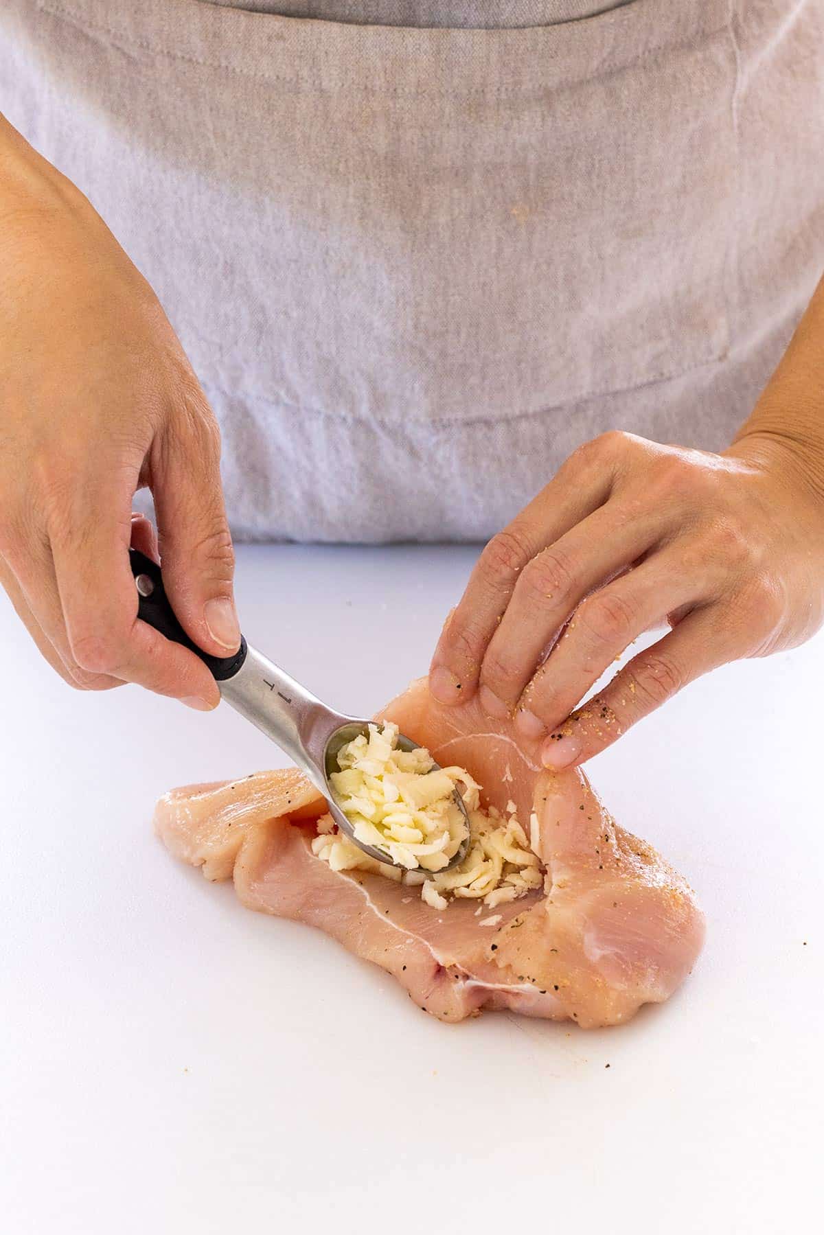 Person using a measuring spoon to place grated mozzarella inside a chicken breast.