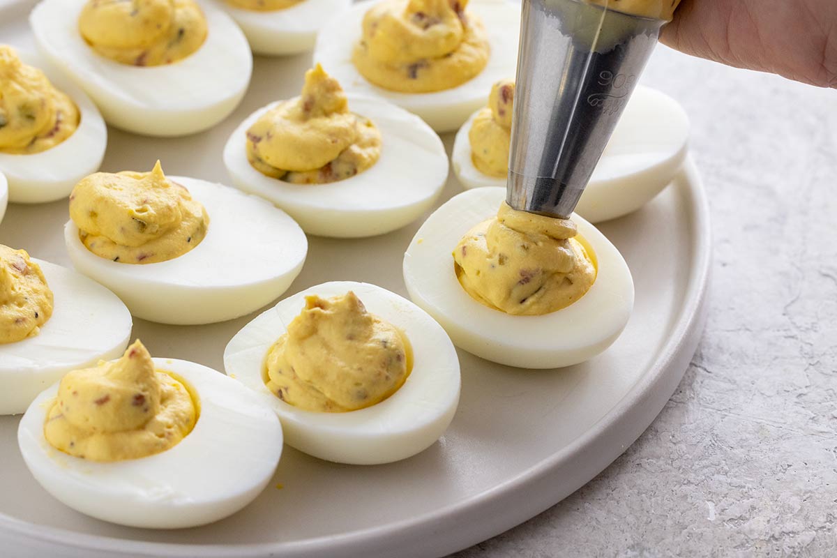 Piping bacon deviled egg filling into hard-boiled eggs.