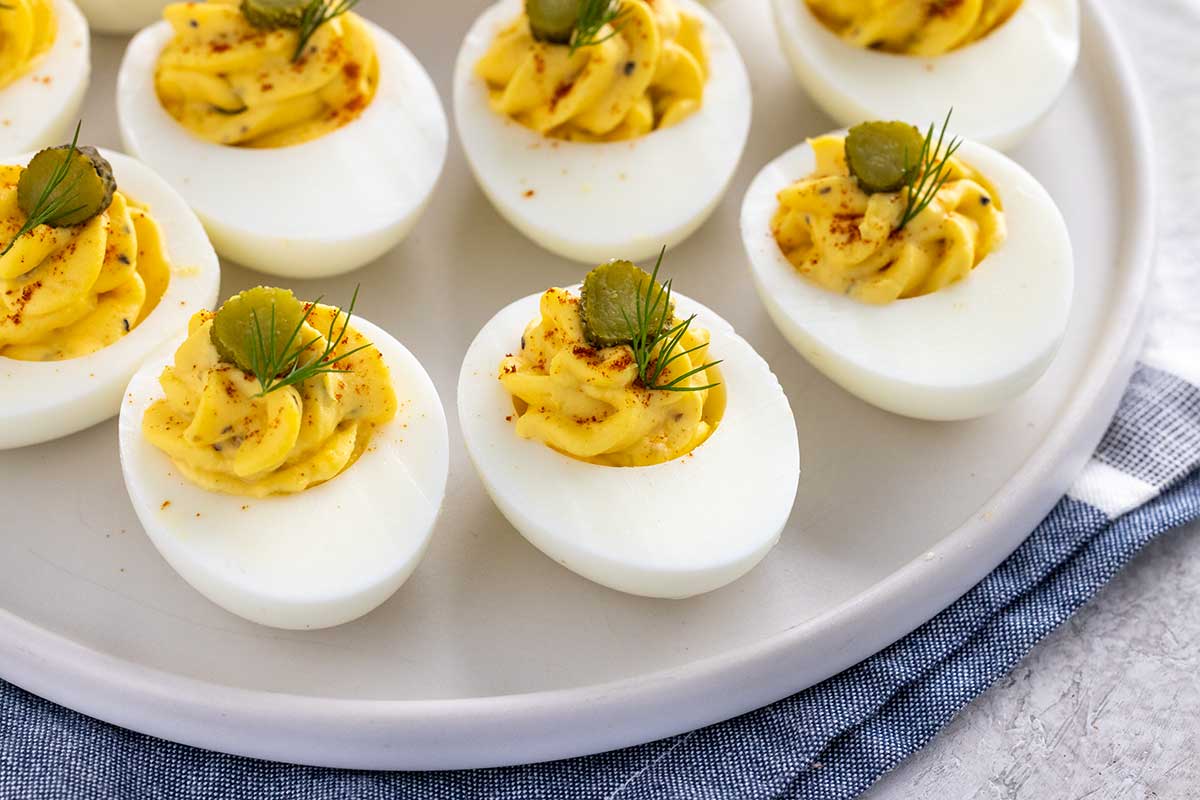 A platter of deviled eggs garnished with dill leaves and sliced gherkins on top.