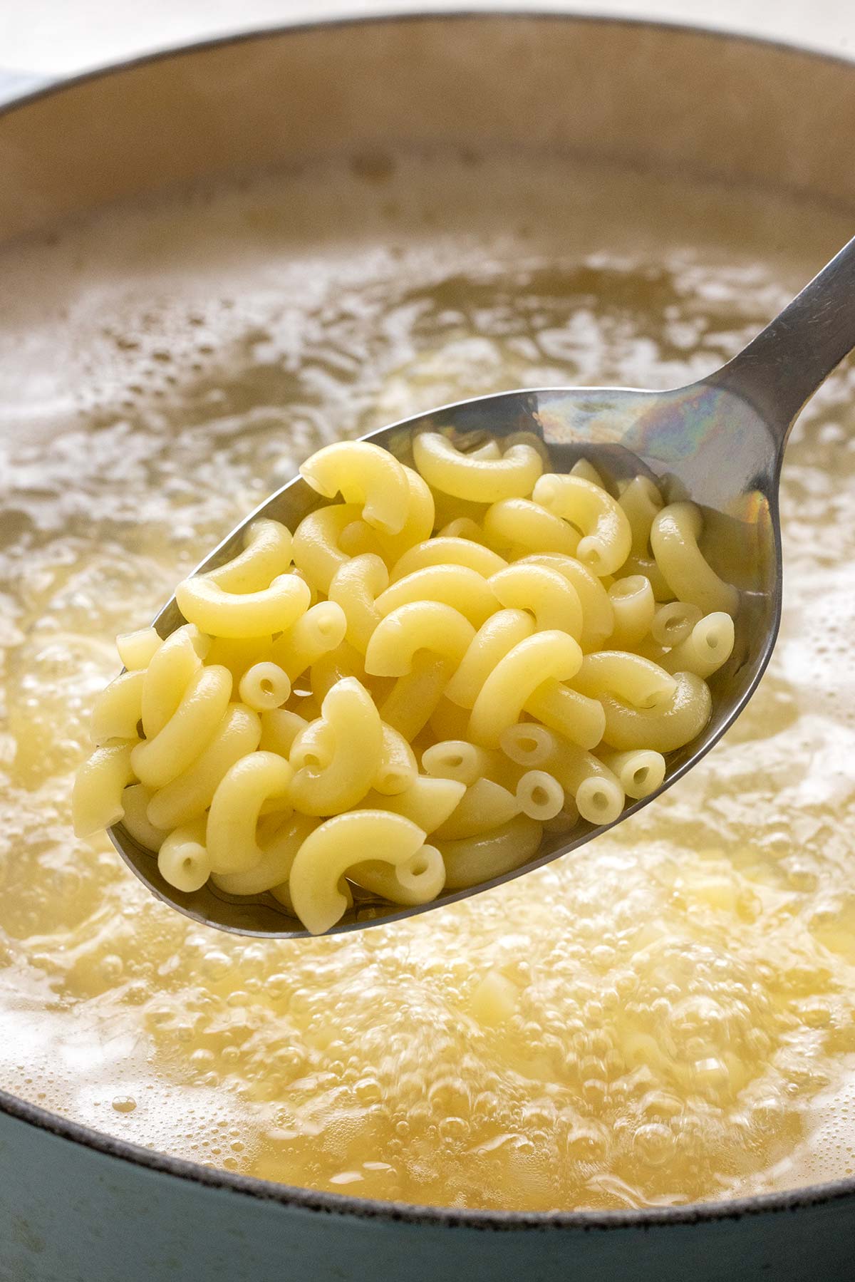Metal spoon lifting cooked macaroni out of a boiling pot of water.