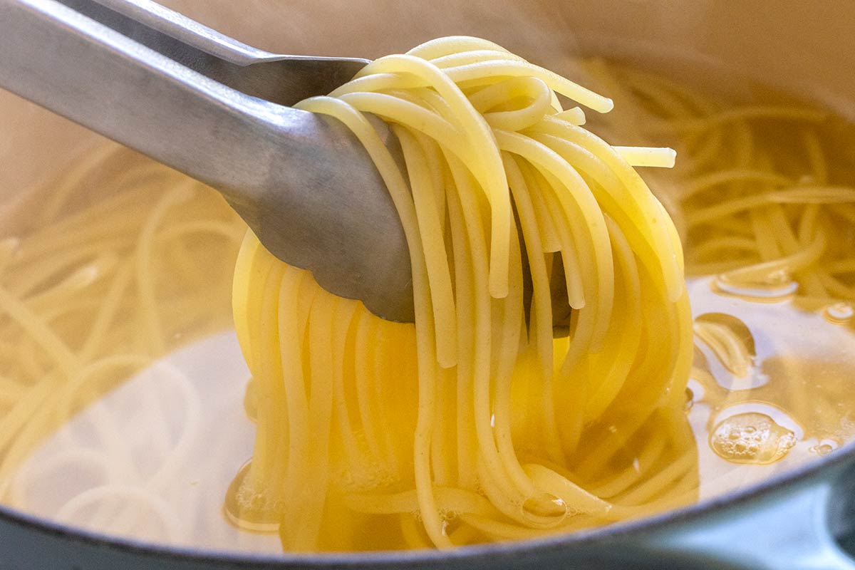 Tongs lifting cooked linguine from a pot of hot water.