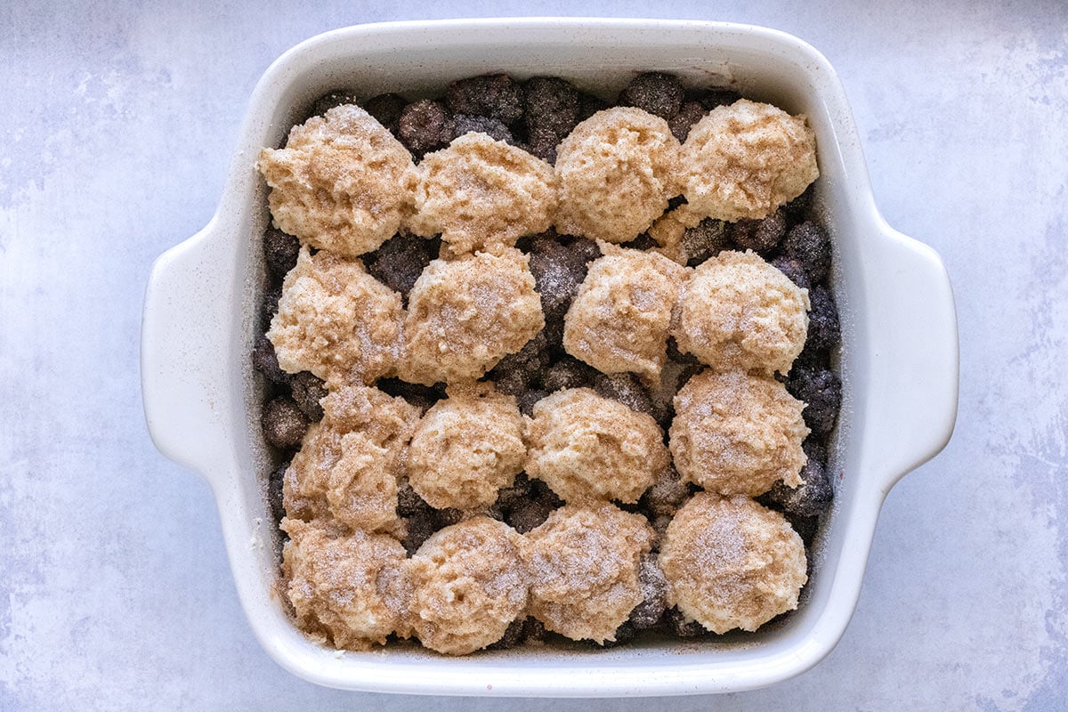 Biscuit dough clumps laid onto blackberries inside a square baking dish.