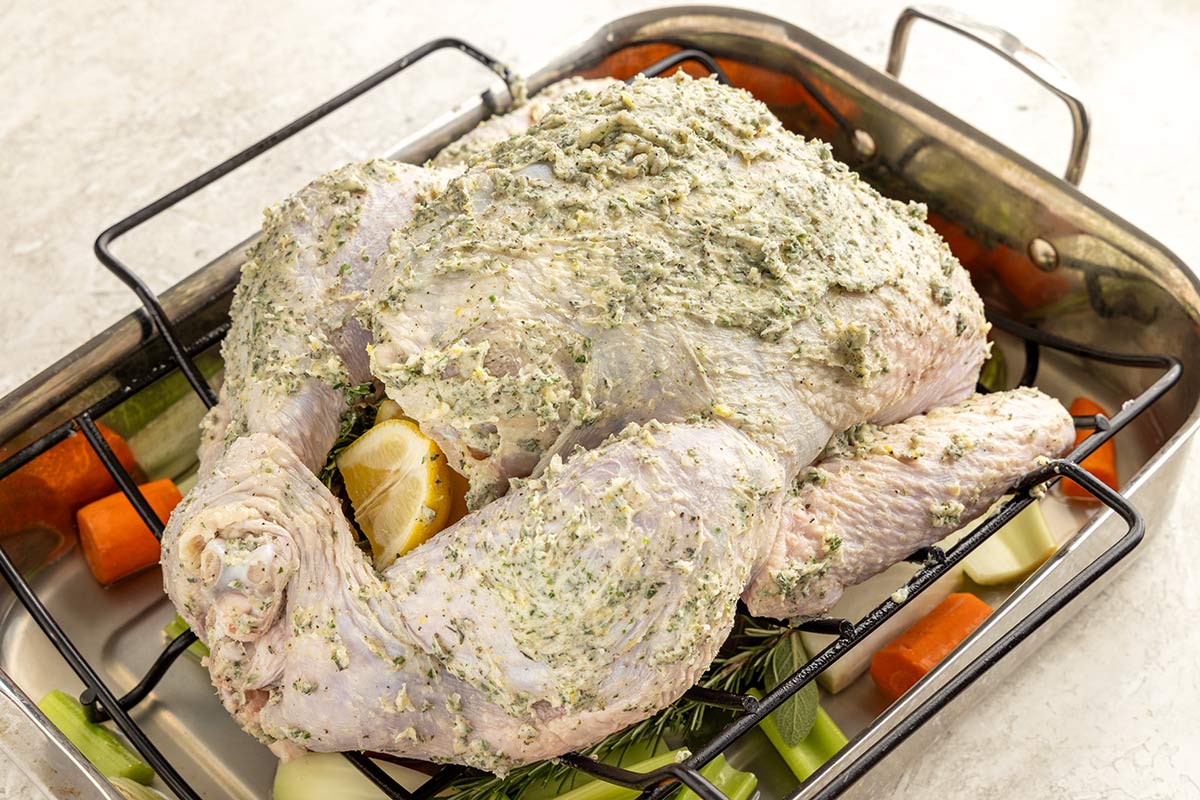 Turkey covered in herb butter placed on a rack and roasting pan with vegetables.