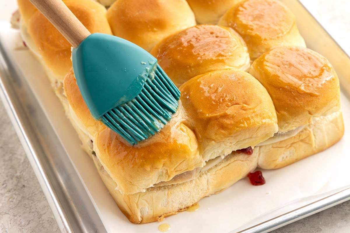 Brush applying melted butter to the top of Hawaiian rolls on a sheet tray.