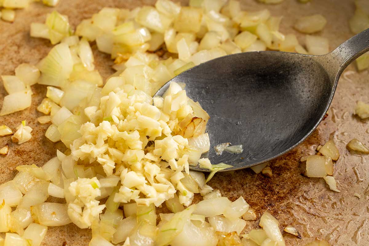 Metal spoon stirring diced yellow onion and minced garlic.