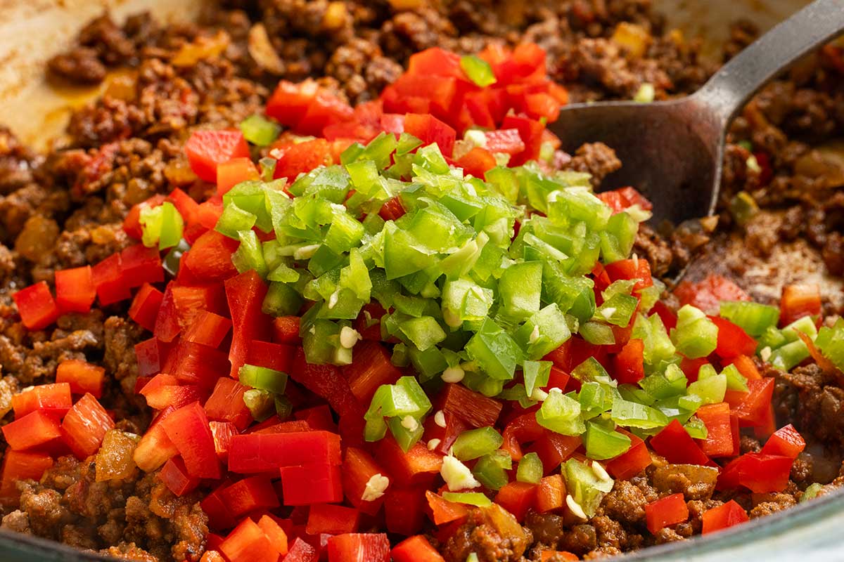 Diced pieces of jalapeno and red bell pepper placed in a pan with ground beef.