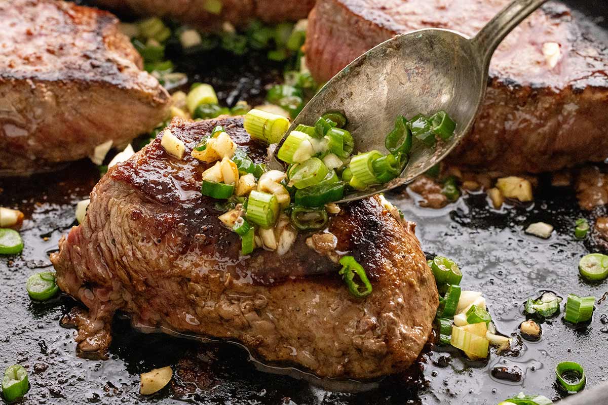 Spoon adding minced garlic and sliced green onions to the top of a steak fillet.