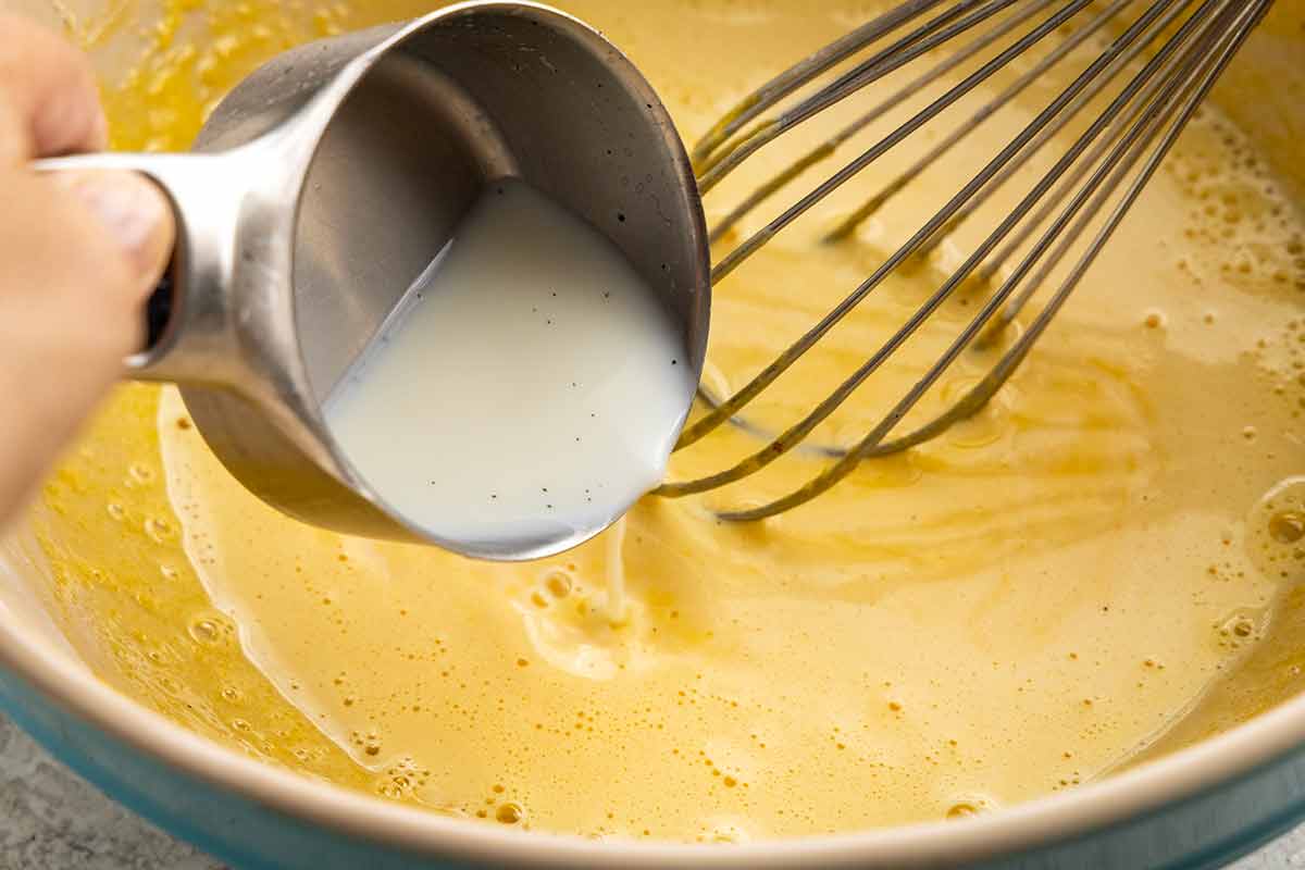 Measuring cup pouring hot milk into a bowl of whisked egg yolks.