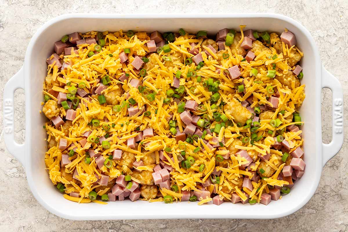 Ham, cheese, tater tots, and green onions placed inside a white casserole dish.