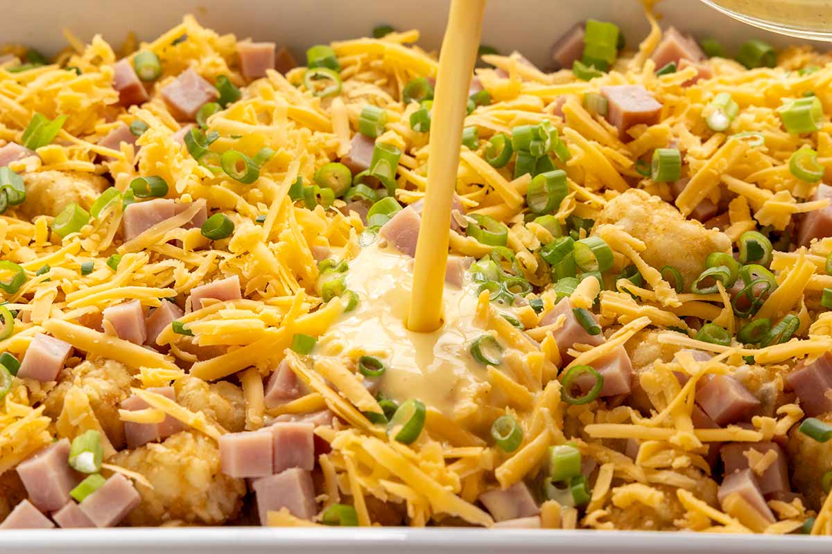 Egg custard being poured over shreds of cheddar, cubes of ham, and frozen tater tots.