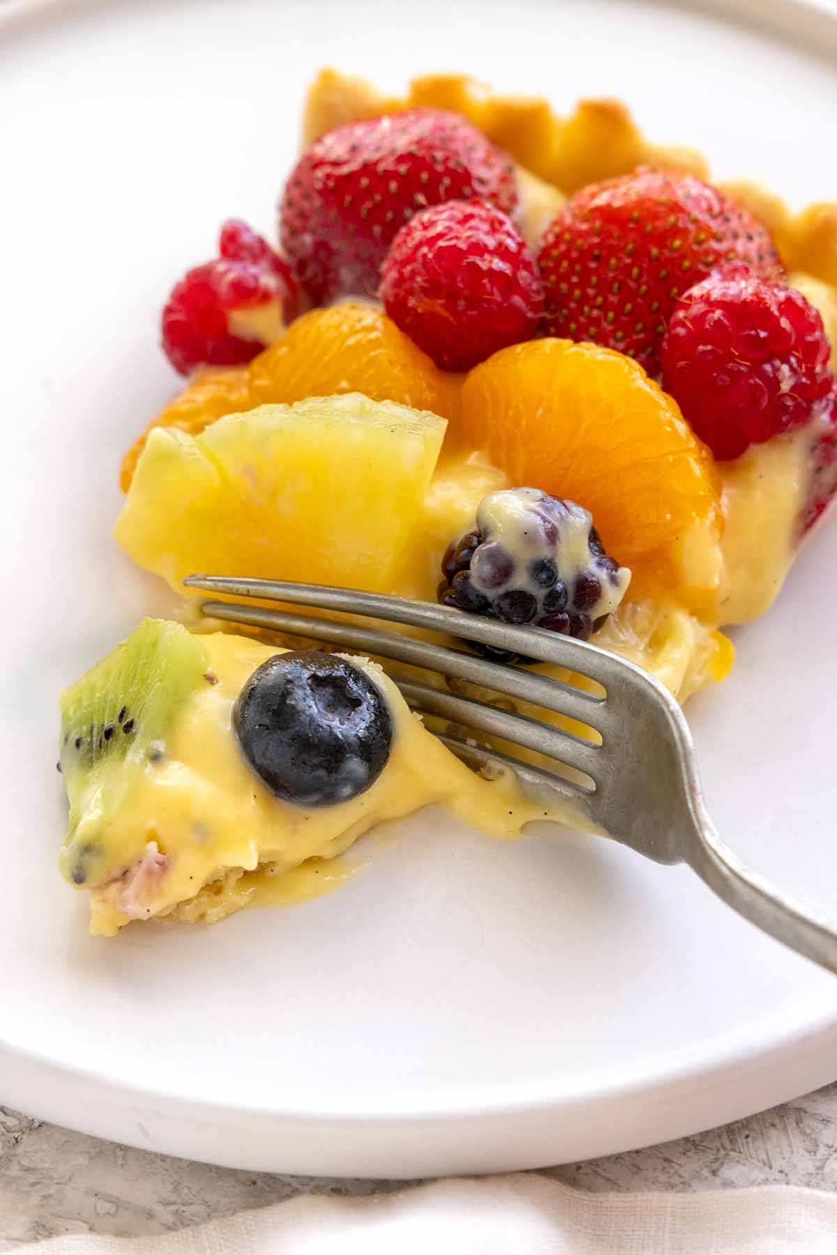Fork cutting into a slice of fruit tart with blueberries, raspberries, and oranges.