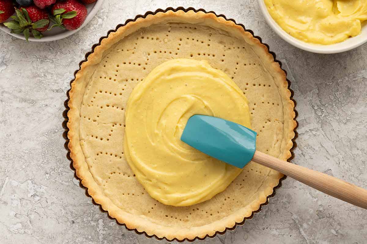 Blue spatula spreading pastry cream into a baked tart crust.
