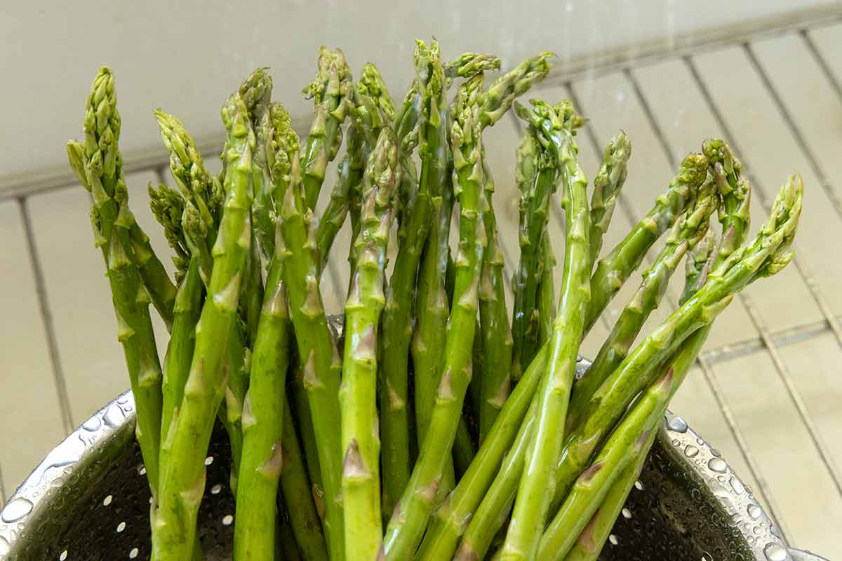 Asparagus spears in a metal colander being rinsed under a kitchen sink.