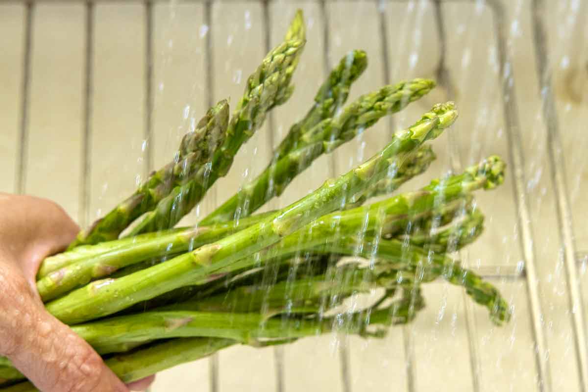 Person holding a bunch of asparagus by the stalks and rinsing them under running water.