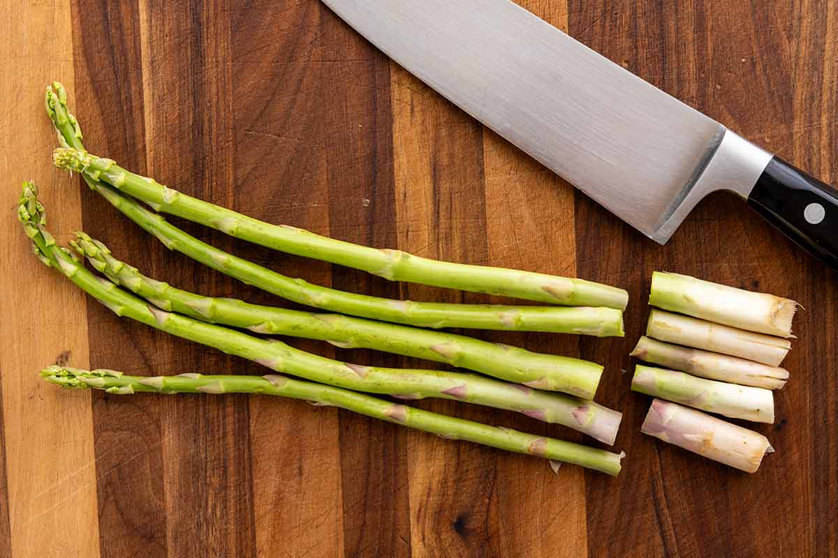 Wooden cutting board with a knife next to asparagus with ends sliced off.