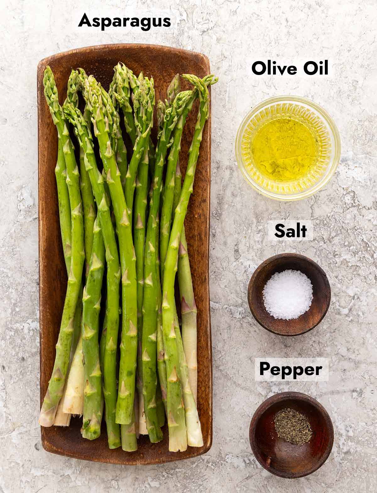 Asparagus spears on a wooden plate next to three small bowls that are filled with olive oil, salt, and pepper.